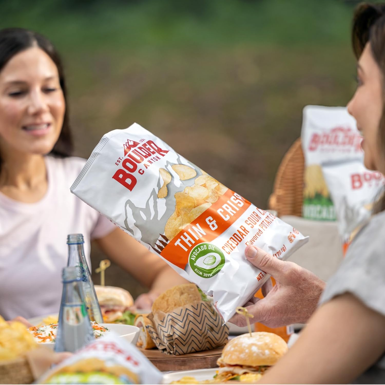 Two people enjoying a meal outdoors with a focus on a bag of Boulder Canyon chips.