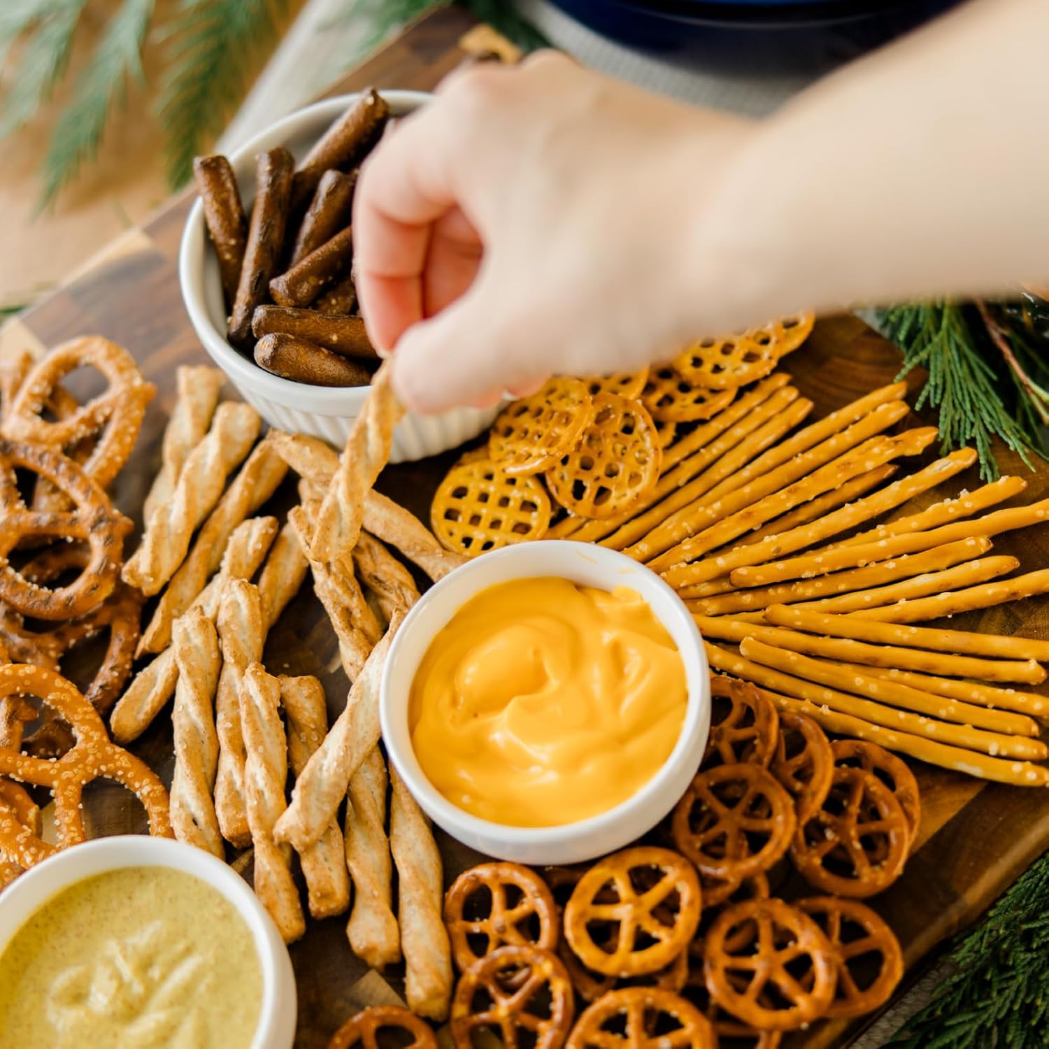 Assorted pretzels and cheese dip on a wooden board with a hand dipping a pretzel.