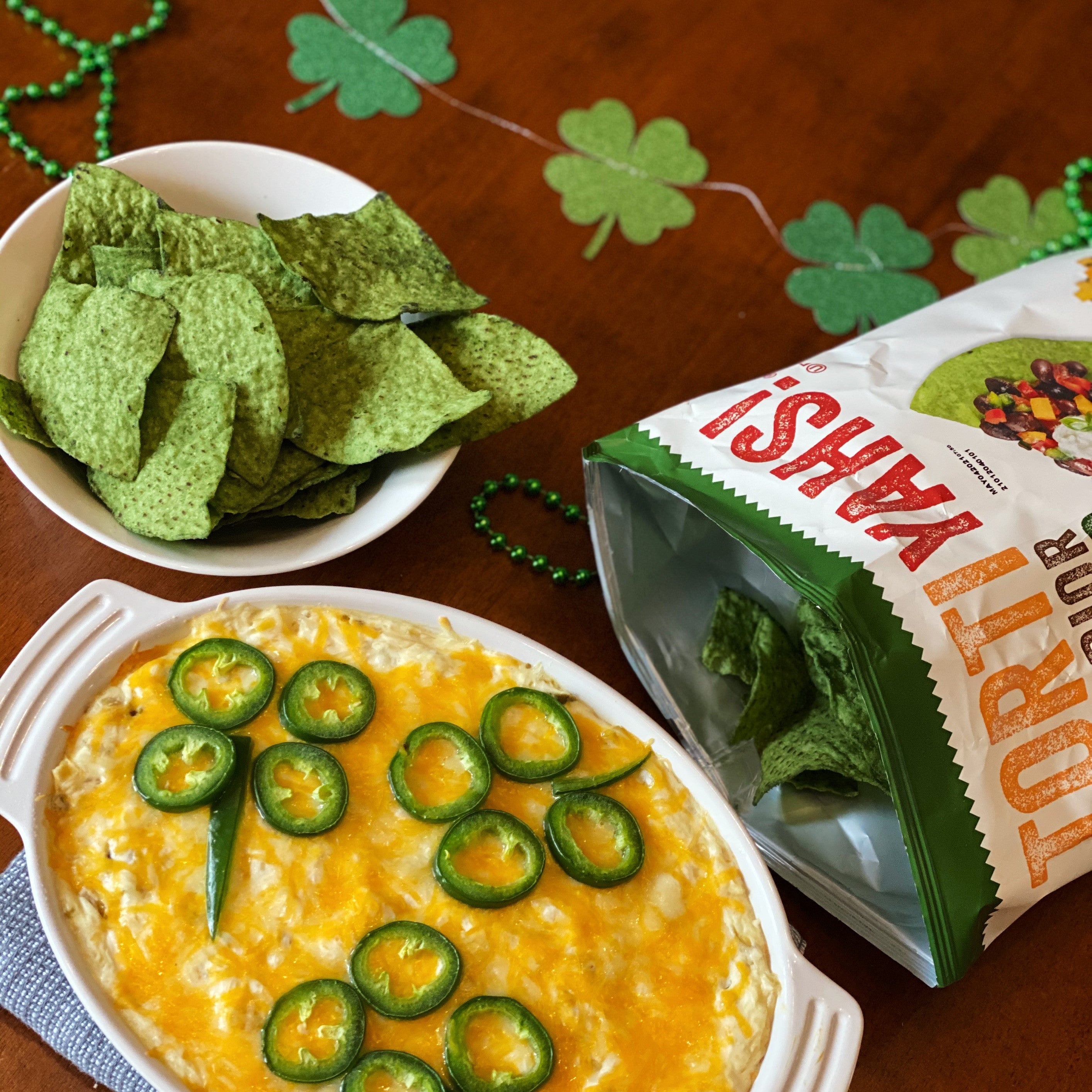 Cheesy dip with jalapeños, tortilla chips, and Yashi tortilla chips packaging on a wooden surface.